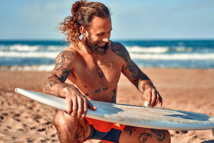 Tattooed surfer waxing his surfboard on the beach